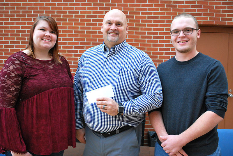 female student in red shirt and male student in black sweater smiling at the camera. between them is a man in a blue checkered dress shirt holding a check.