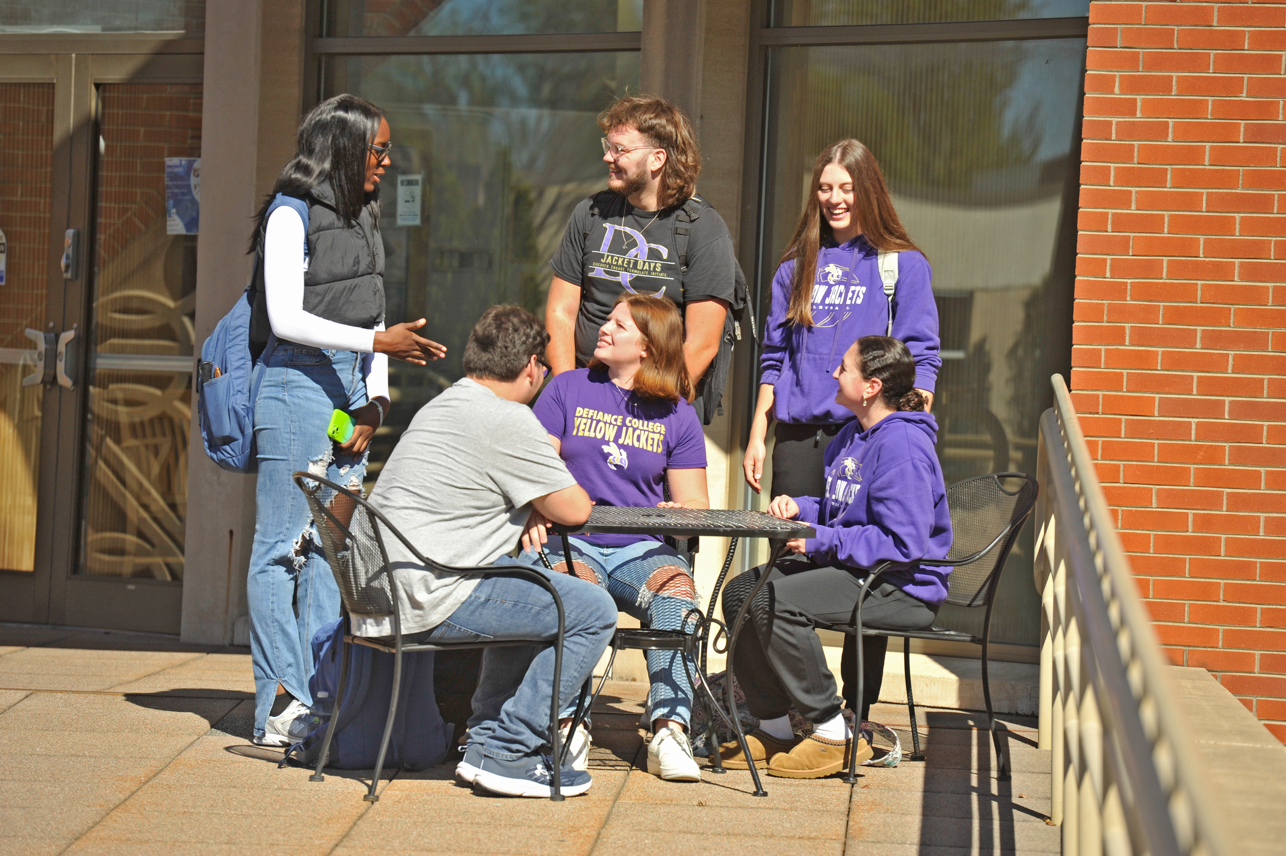 Four female students and two male students outside of a brick building with tall glass windows talking and laughing